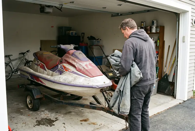 Owner holding a folded storage cover, looking down at a jet ski with chalky, peeling plastics in a garage
